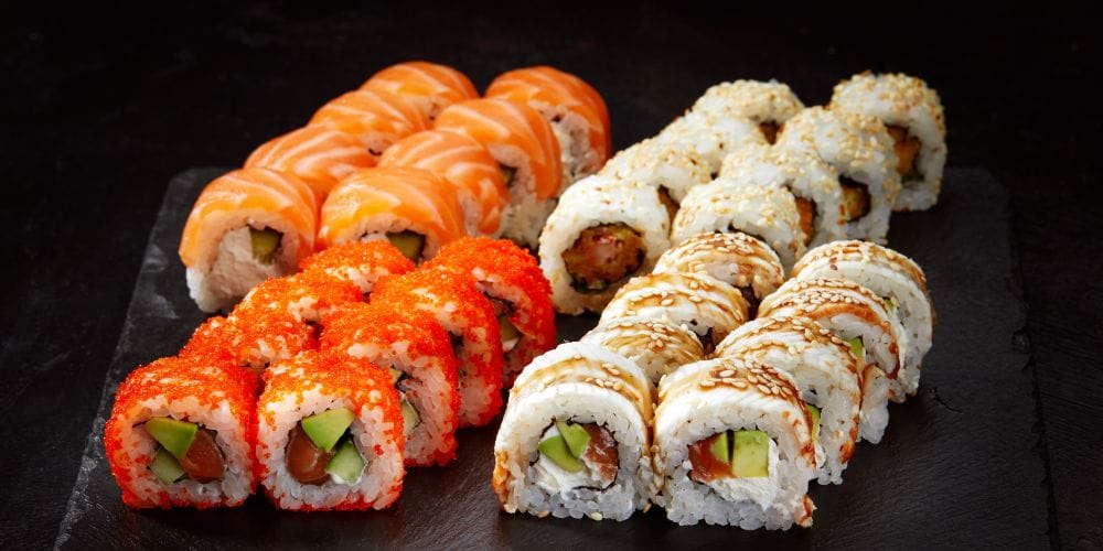 A variety of freshly prepared sushi rolls, including salmon, tobiko, and eel rolls, served on a black slate board