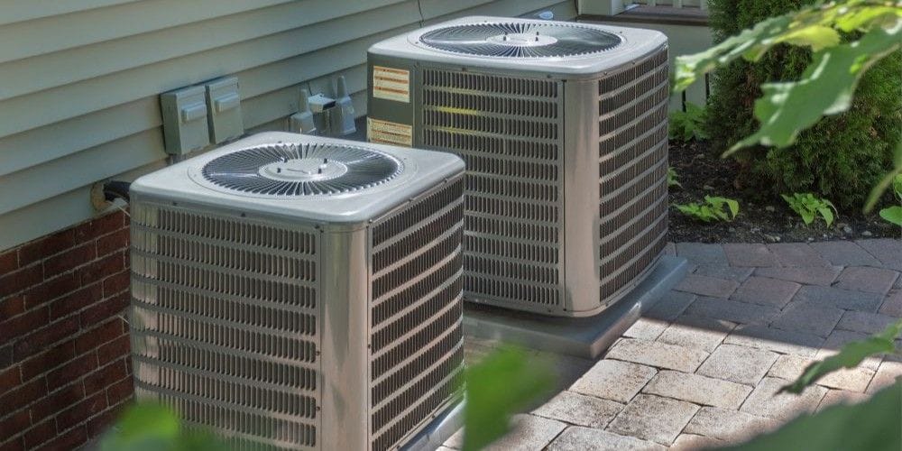 Two outdoor residential air conditioning condenser units installed outside a home on a stone patio