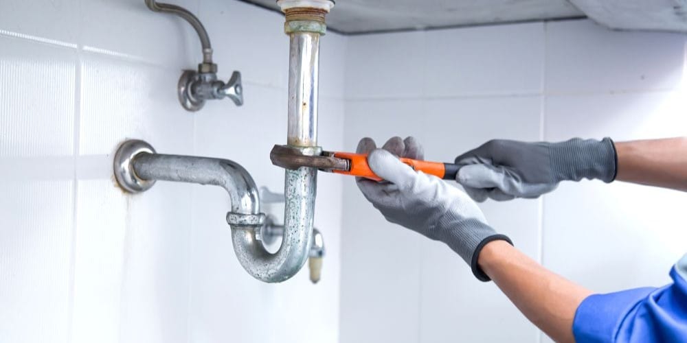 Plumber in protective gloves using a pipe wrench to repair a metal sink drain pipe against a white tiled wall.