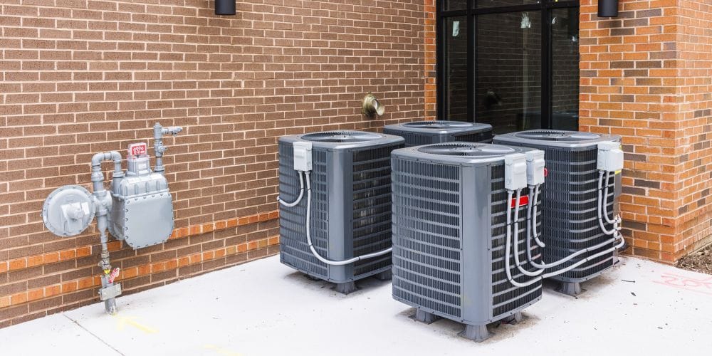 Row of outdoor air conditioning condenser units installed on a concrete pad outside a brick building