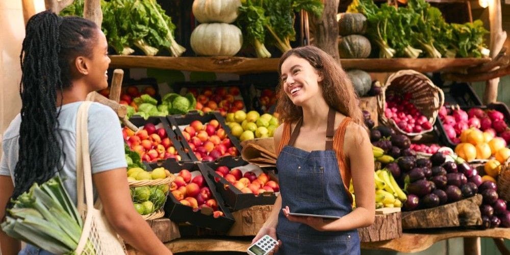 A smiling worker in a denim apron assisting a customer shopping for fresh vegetables and fruit at an open-air community market stall.